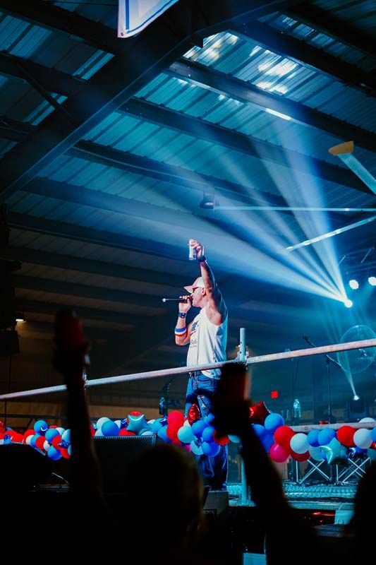 Performer on stage with raised arm, blue spotlights, and red-white balloons in a crowded indoor venue