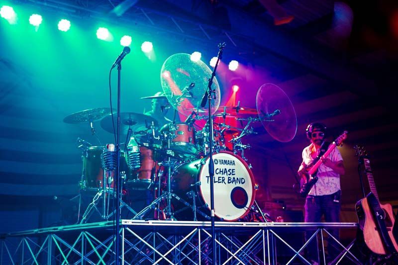 Rock band performing onstage under colorful lights, with a drummer, guitarist, and large drum kit.