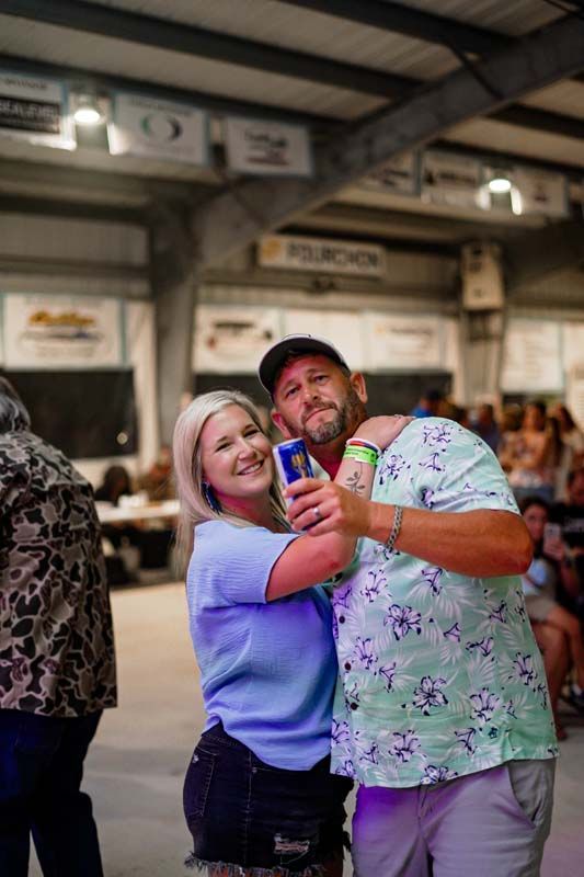 Two people smiling and posing together indoors at a lively event, holding a can and camera.