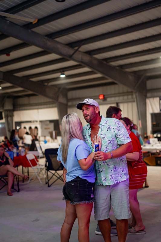 Two people talking and holding drinks in an indoor event hall with folding chairs and a crowd in the background