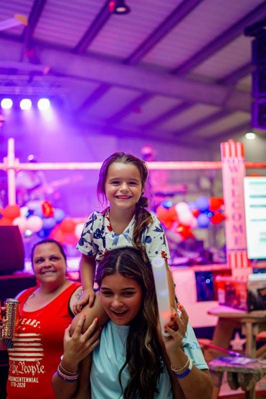 Smiling child sitting on an adult’s shoulders at a colorful indoor event with purple lights and balloons