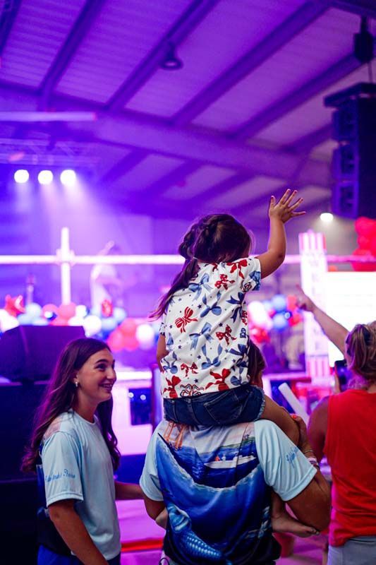 Child on adult’s shoulders at a concert, waving under purple stage lights