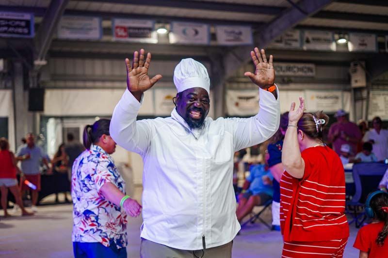 Smiling chef in a white coat raises both hands in a busy indoor hall while people dance around him