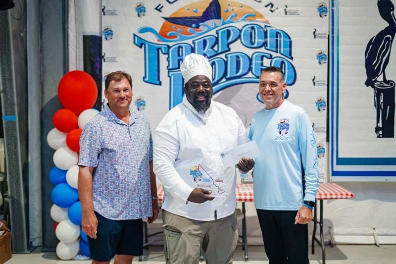 Three men posing in front of a “Tarpon Rodeo” sign at an event booth