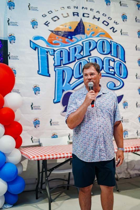 Man speaking into a microphone in front of a Tarpon Rodeo event banner and red, white, and blue balloons
