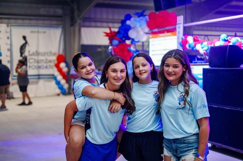 Four teammates in light blue shirts smiling and posing indoors beside red, white, and blue balloons.
