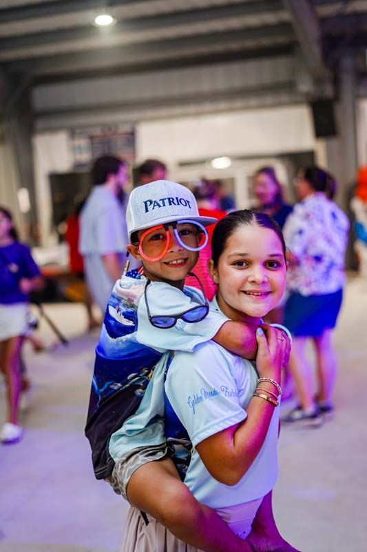 Two children posing at an indoor event, one giving the other a piggyback ride, both smiling.