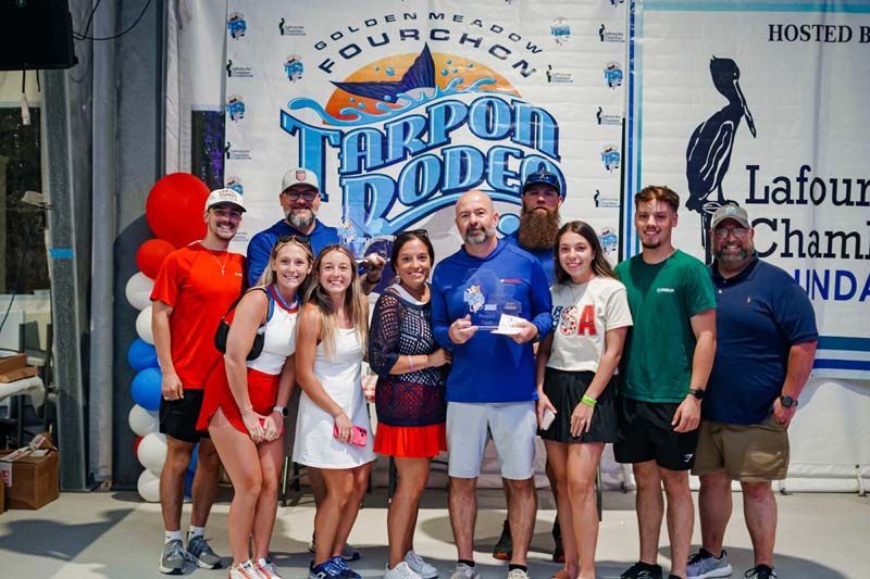 Group of people posing in front of a Tarpon Rodeo sign, with balloons and event shirts, smiling outdoors