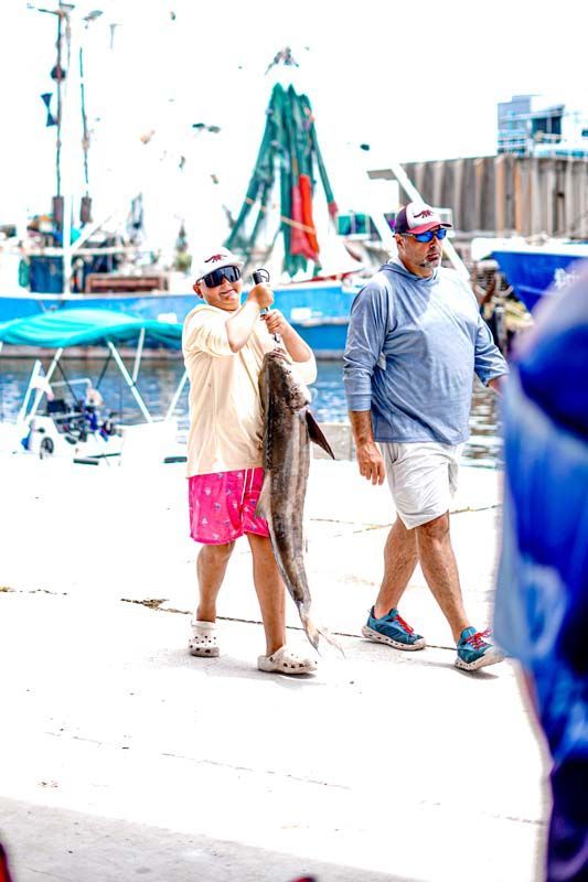 Two people walking by a marina with boats and bright sunlight.
