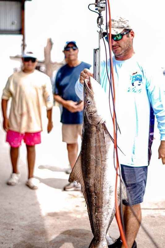 Man holding a large fish on a hook, with two people standing in the background near a dock.