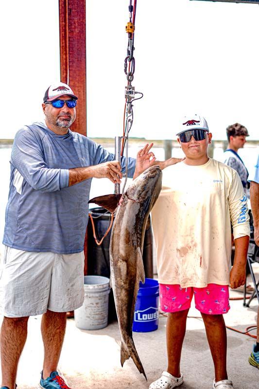 Two people holding up a large fish on a boat deck, with fishing gear and the ocean in the background.