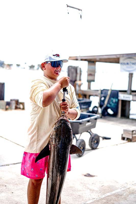 Person holding a large fish at a marina, wearing a cap, sunglasses, tan shirt, and bright pink shorts