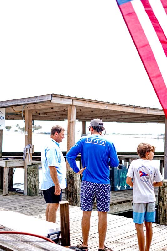 People standing on a dock under a wooden shelter near the water, with a red banner at the right.