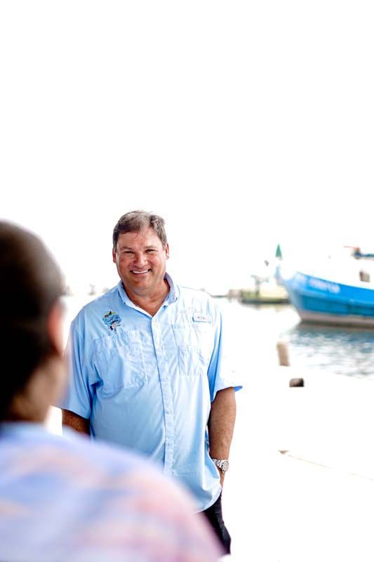 Man in a light blue shirt standing by the water near a docked boat, smiling outdoors