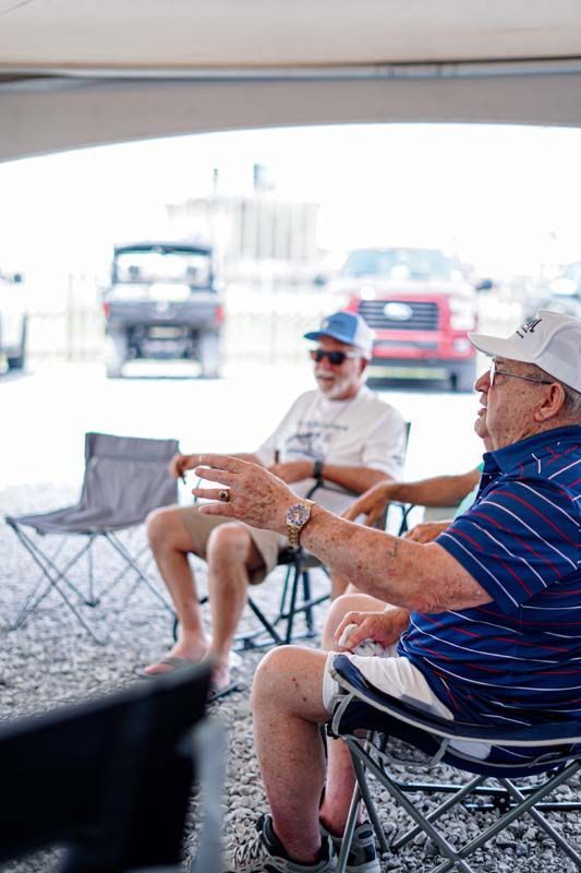 Two men sitting in folding chairs under a canopy, chatting at an outdoor event with cars in the background