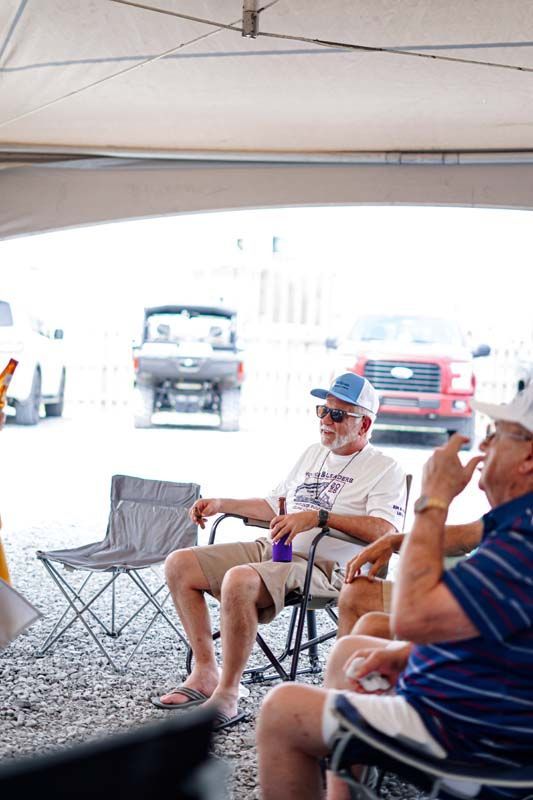People sitting in folding chairs under a canopy at an outdoor gathering, with parked cars in the background