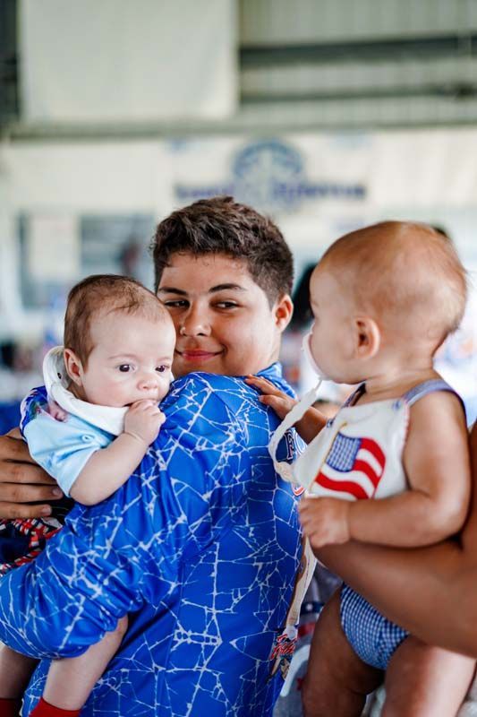 Person holding two infants in blue outfits, one with an American flag shirt, indoors.