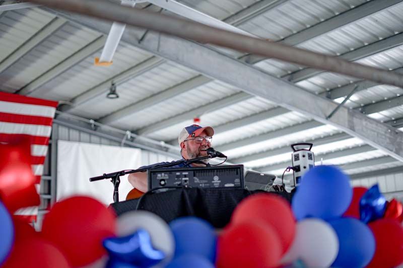 Person speaking at a podium in a hangar, framed by red, white, and blue balloons and an American flag.
