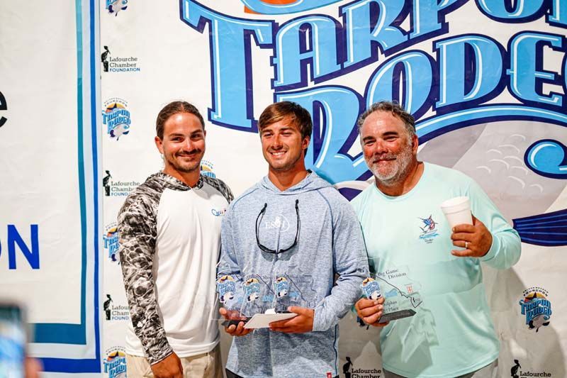 Three men are standing next to each other holding trophies in front of a tarp rode sign.