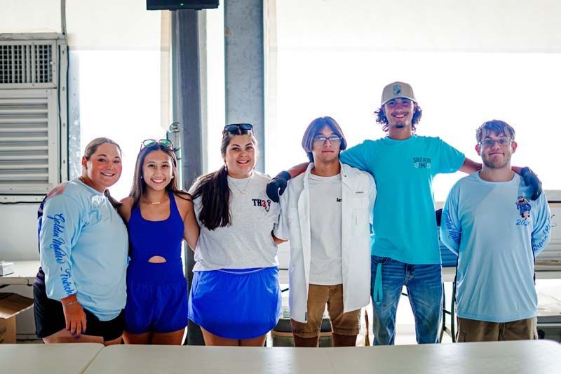 Six people standing together indoors, smiling, with windows and an air conditioner behind them