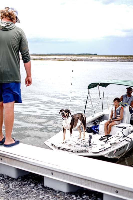 Man on dock facing a small boat with two people and a dog on calm water