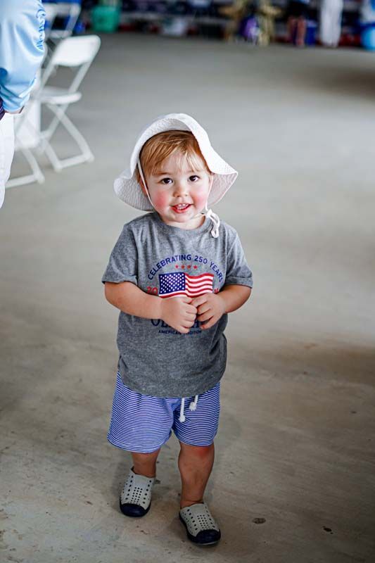 Toddler standing indoors, holding a small flag and wearing a white sun hat, gray shirt, and blue shorts