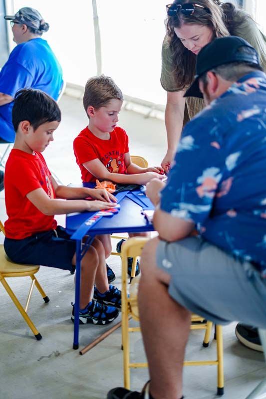 Two children in red shirts play a tabletop game with adults nearby in a bright room