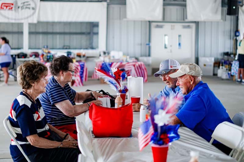 People seated at a long table in a bright hall, holding red-white-blue flags and cups, socializing.