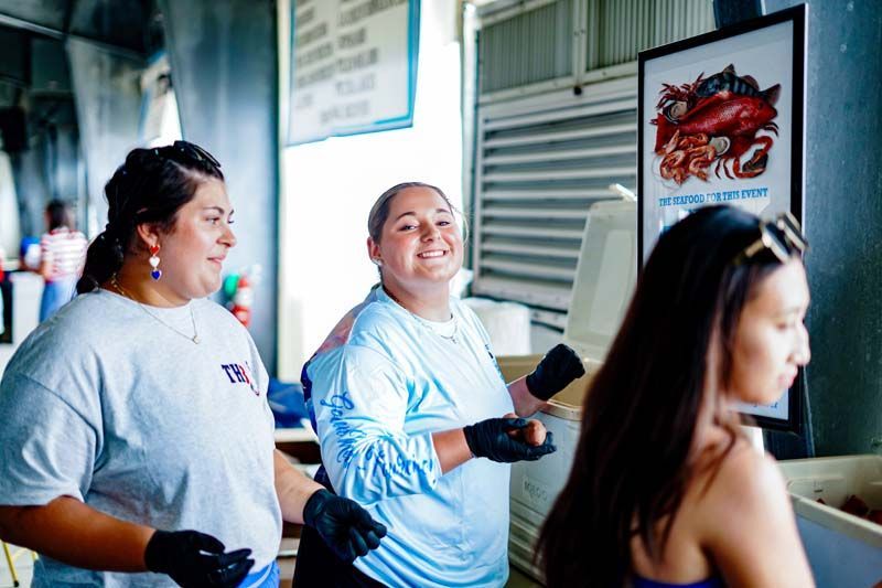 Three people in light blue shirts smiling indoors near a counter and a framed lobster poster.