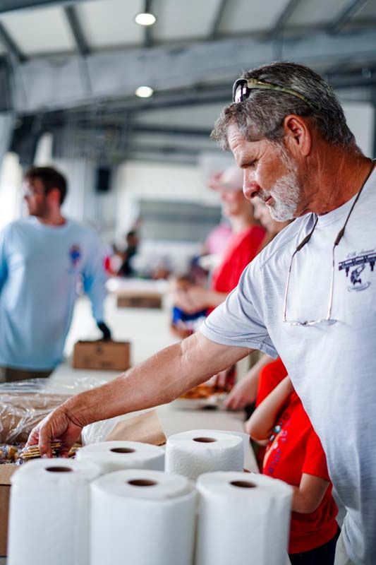 Man in white shirt stacks toilet paper rolls at a table in a bright indoor space