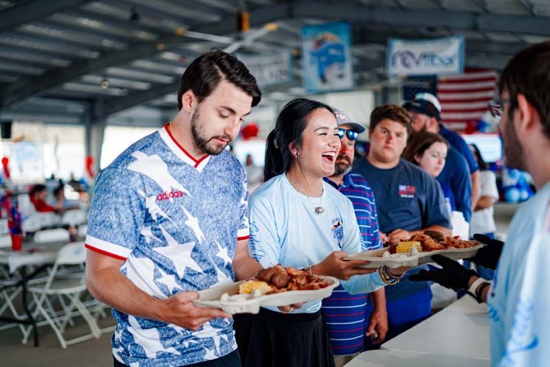 People serving food at a patriotic-themed event under a covered pavilion