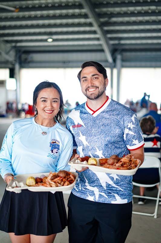 Two people holding trays of food at an indoor event, both wearing team jerseys and smiling.