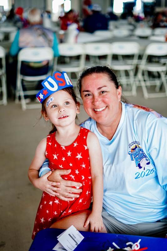 Smiling pair seated at event table, wearing red and light blue patriotic outfits with stars and a USA headband.
