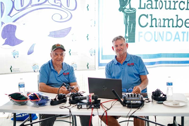 Two men in blue shirts sit at a table with audio gear and a laptop at an event booth.
