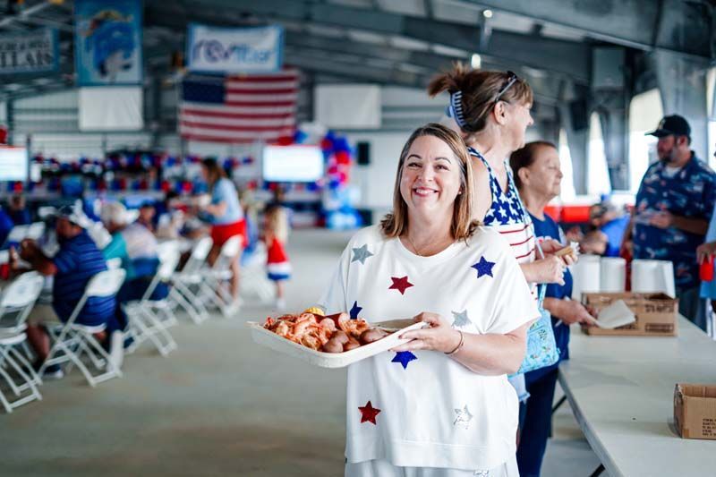 Volunteer handing out food at an indoor community event, with U.S. flags and seated attendees in the background