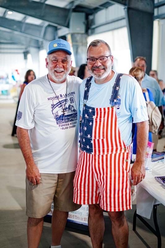 Two smiling men in casual clothes at an indoor event, one wearing red-and-white striped overalls and an American flag shirt