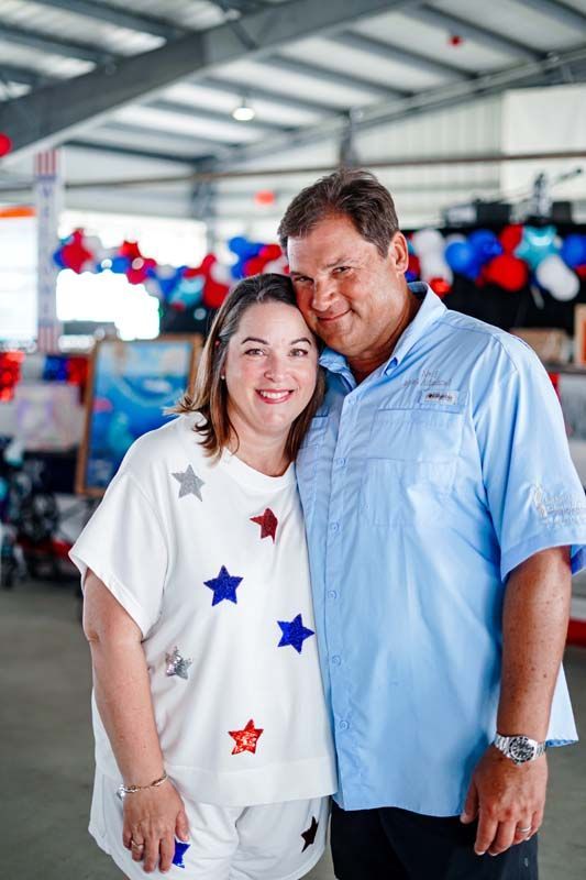 Two people smiling indoors at an event, with red, white, and blue balloons in the background.
