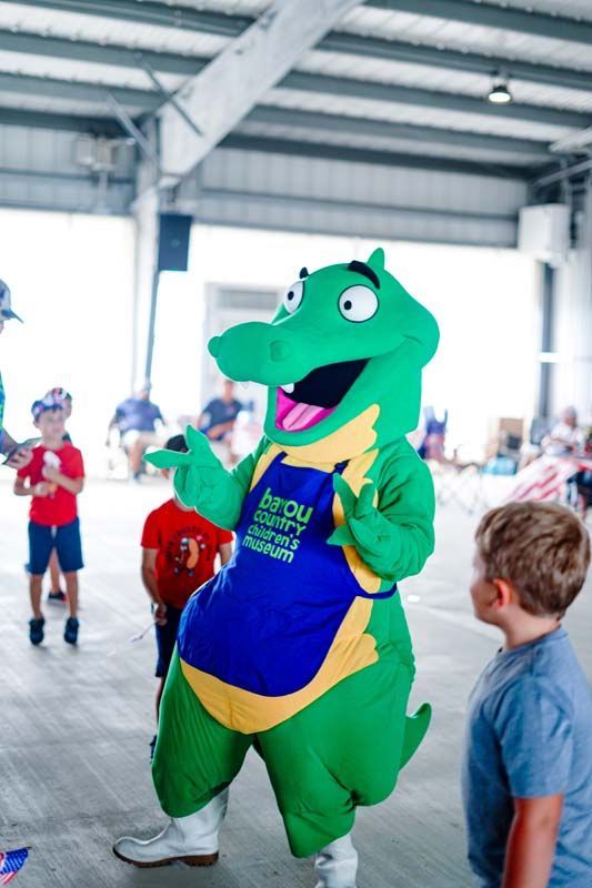Green dragon mascot in a blue apron waving at children inside a bright indoor event space