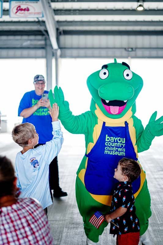 People high-five a green bayou mascot in a blue and yellow shirt inside a covered venue.
