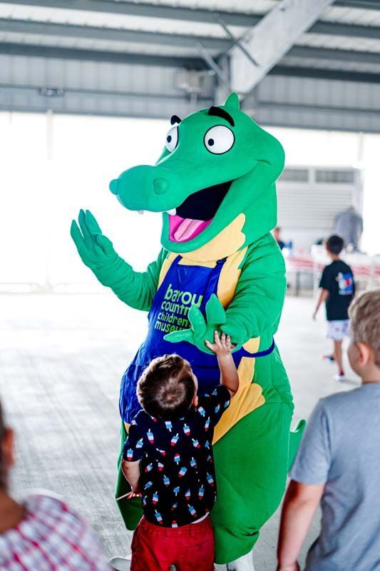 Green alligator mascot waving at children indoors under a covered pavilion