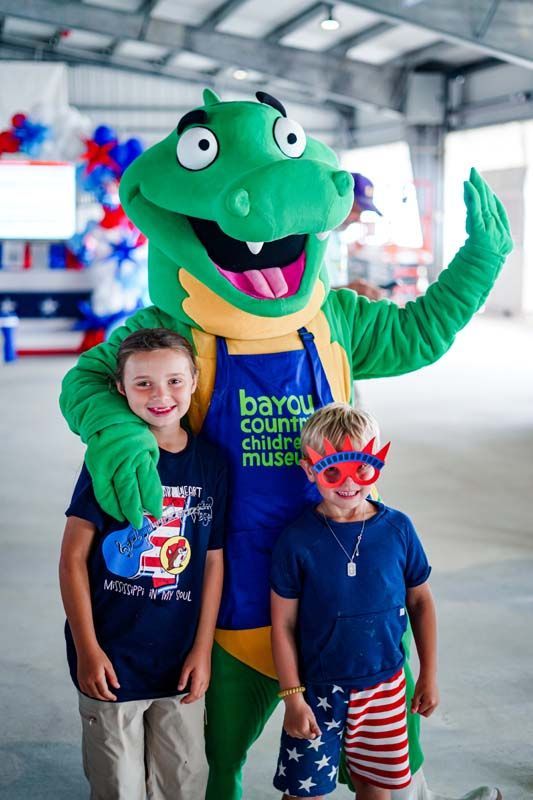 Costumed dinosaur mascot posing with two children in a museum, one child wearing star-spangled shorts and red glasses