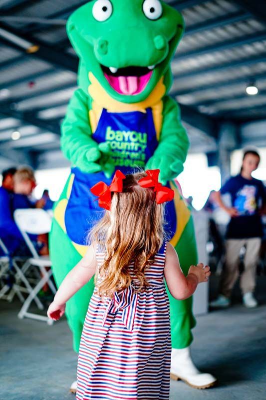 Green Bayou mascot greeting a child indoors, with a red bow in the girl’s hair