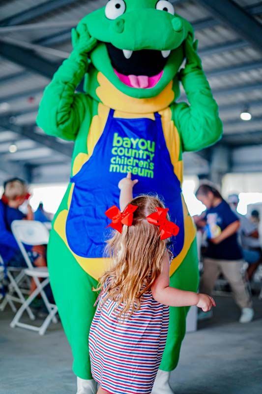 Green dinosaur mascot greeting a child with red bow in an indoor event space