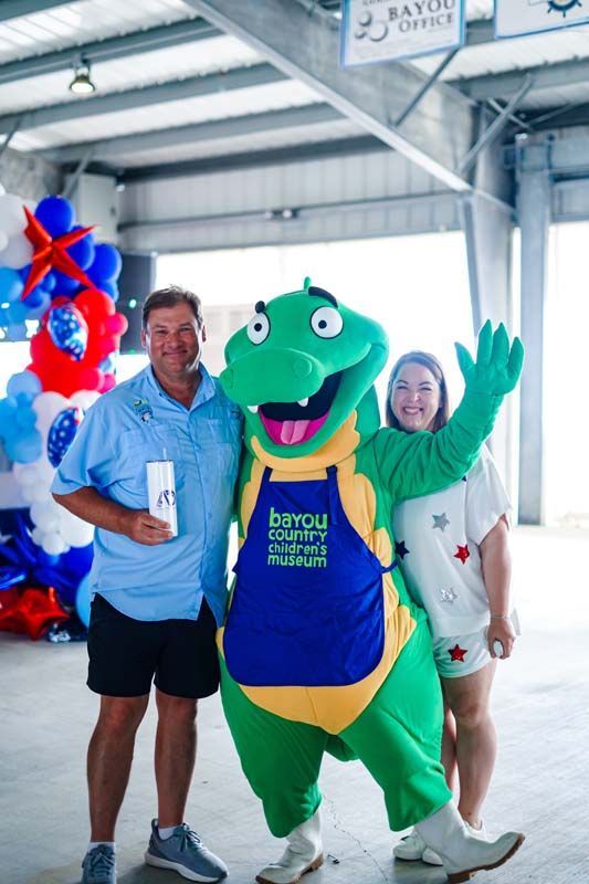 Man in blue shirt and green dragon mascot posing under a pavilion with balloons and a Bayou festival banner