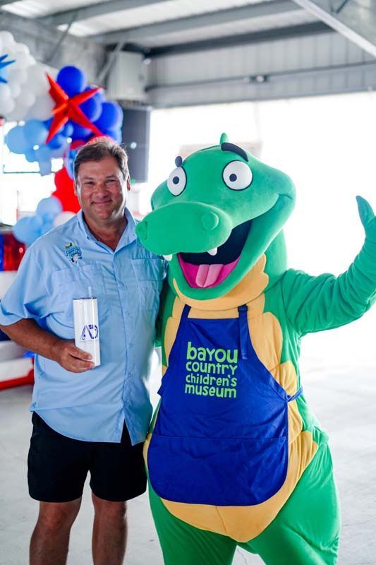 Man posing with a teal crocodile mascot in a blue apron at an indoor event, holding a drink cup