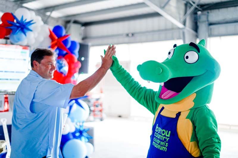 Man high-fives a green mascot in a decorated indoor event space with red, white, and blue balloons