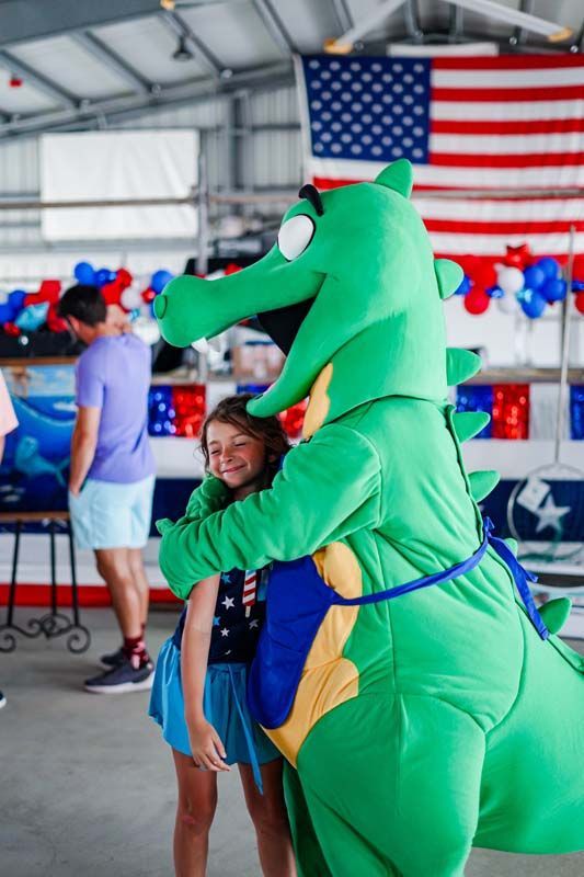 Person in an alligator costume hugging a child at an indoor event with an American flag backdrop