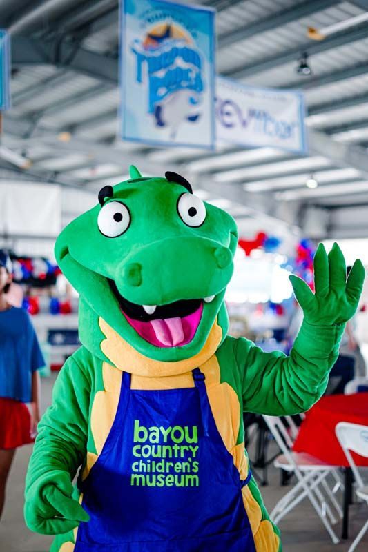 Green dinosaur mascot waving, wearing a yellow shirt and blue apron at Bayou Country Children's Museum.