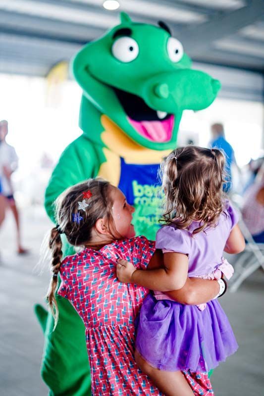 Mascot in green costume hugging two small children, one in purple dress, at an indoor event