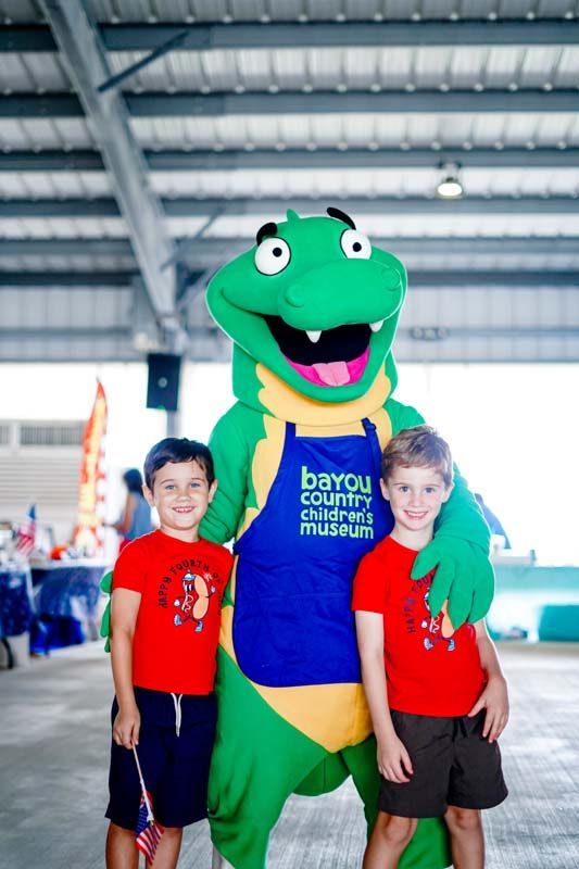 Green mascot in blue shirt stands with two smiling children in red shirts at an indoor event